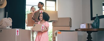Two adults and a little girl walk into a home while holding boxes