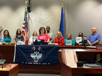 Coralys Santana, a woman with curly hair and glasses and wearing a pink shirt, stands at a podium. In front of her is a UConn HUSKY banner